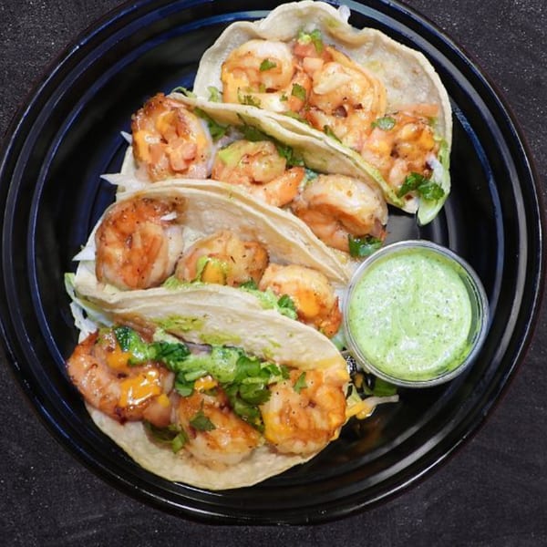 Three shrimp tacos with cilantro, cheese, and a side of green sauce in a black bowl.