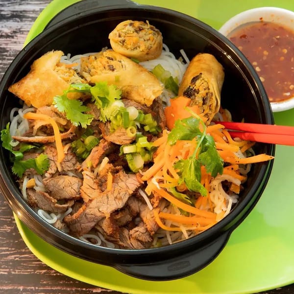 Pho Go's vermicelli bowl with sliced beef, spring rolls, carrots, and herbs, with a side of sauce.