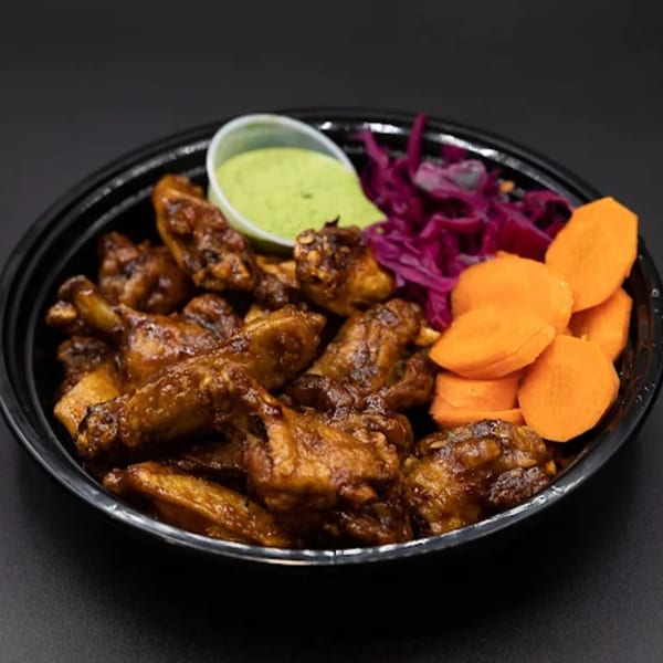 A black bowl filled with glazed chicken wings, red cabbage, sliced carrots, and a side of green sauce.