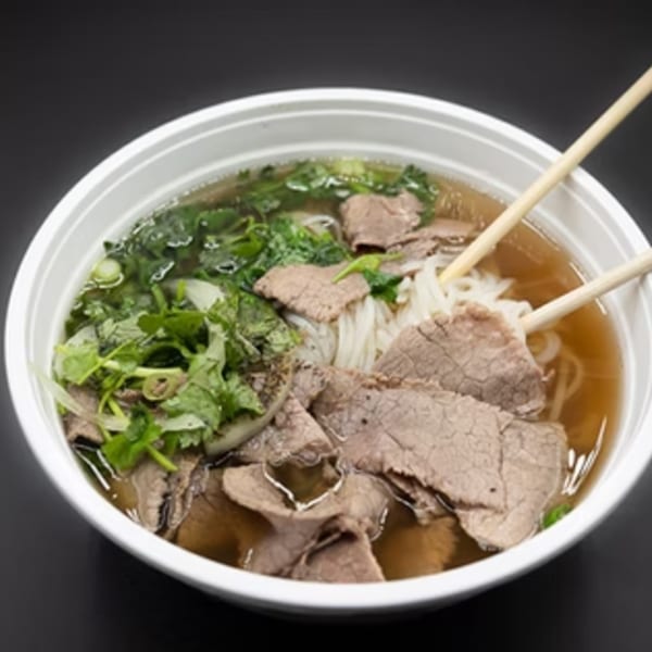 A white bowl of Pho Go pho with beef, noodles, broth, and fresh herbs, with chopsticks.